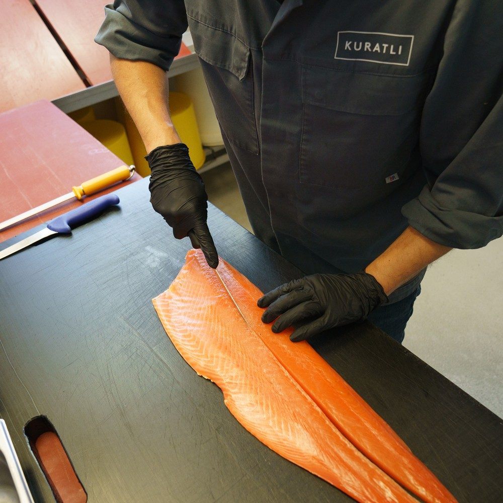 Un homme en vêtement de travail et portant des gants noirs coupe un filet de saumon avec un long couteau fin.