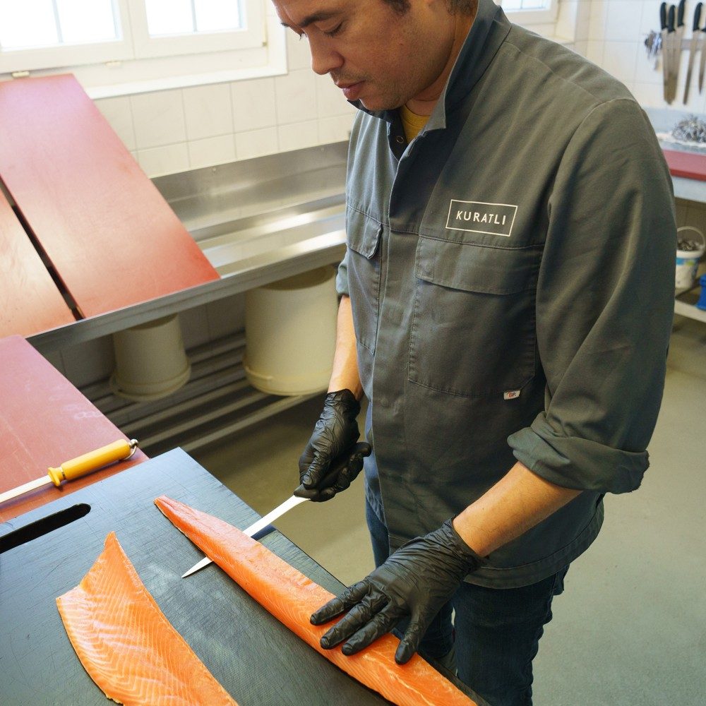 Un homme en vêtement de travail et portant des gants noirs coupe un filet de saumon avec un long couteau fin.