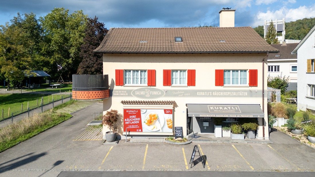 Prise de vue extérieure d’une maison à la façade jaune clair, avec un toit brun et des volets rouges. Sur le store au rez-de-chaussée figure le logo «Kuratli».