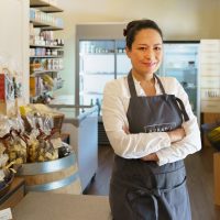 Une femme se trouve dans un magasin. Elle porte un tablier et un chemisier et se tient les bras croisés. Derrière elle sont présentés diverses délicatesses et spécialités dans des rayons, sur un fût de vin et dans un congélateur. A côté d’elle se trouve une caisse et un appareil de paiement électronique.