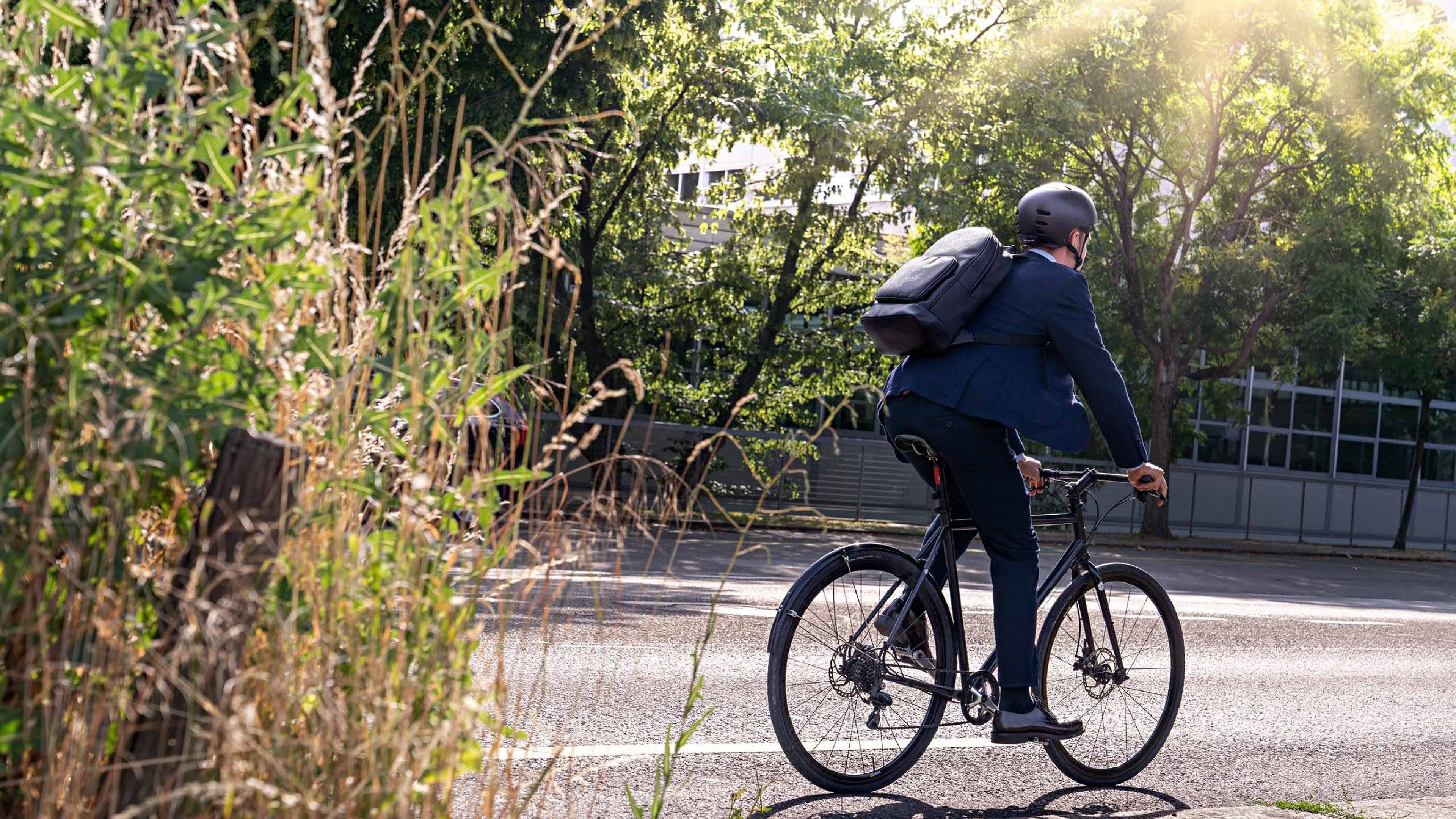 Person in Anzug fährt auf einem Fahrrad entlang einer Strasse, trägt einen Fahrradhelm und einen Rucksack. Im Vordergrund sind hohe Gräser, im Hintergrund Bäume und ein Zaun, beleuchtet von warmem Sonnenlicht.