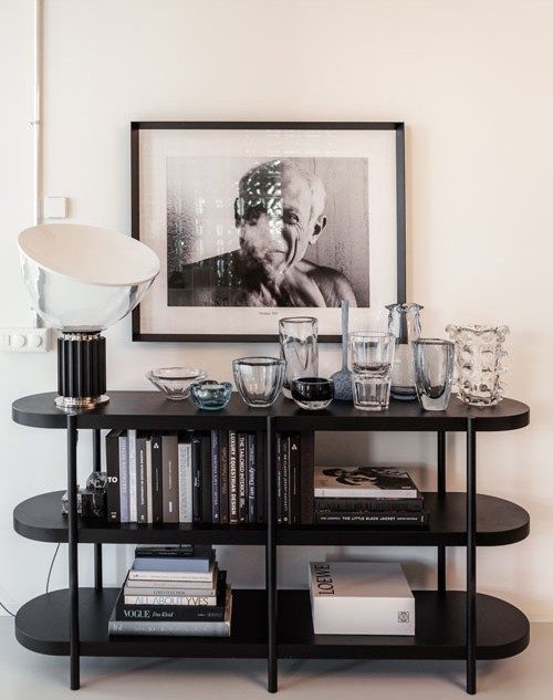 Uno scaffale nero rifornito di libri, eleganti accessori per la casa e una selezione di oggetti in vetro. Sopra lo scaffale è appesa la foto di un uomo in bianco e nero. Un’elegante lampada da tavolo completa l’allestimento sofisticato.