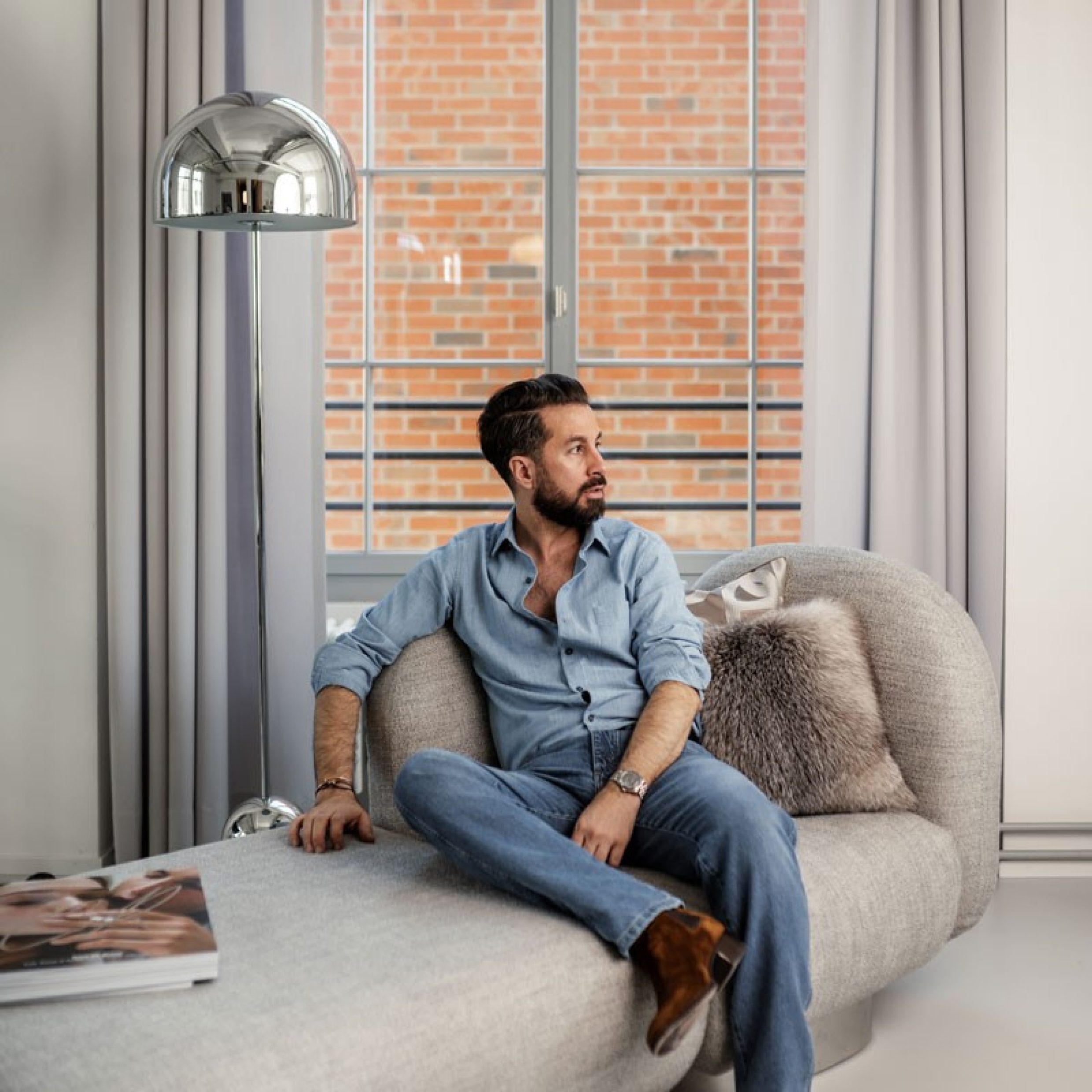 Un uomo dai capelli scuri con camicia in jeans, jeans e stivali in camoscio è seduto su un divano beige e guarda in modo riflessivo di lato. La stanza è arredata con stile, con elementi che si combinano perfettamente.