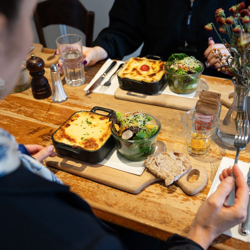 Due persone siedono a un tavolo di legno rustico in un bistrot e mangiano insieme. Sul tavolo ci sono padelline da forno con pietanze gratinate, insalate fresche in ciotole di vetro, fette di pane su taglieri di legno, bicchieri d’acqua e posate. Macinapepe e macinasale, una caraffa e una piccola composizione floreale completano la tavola. 