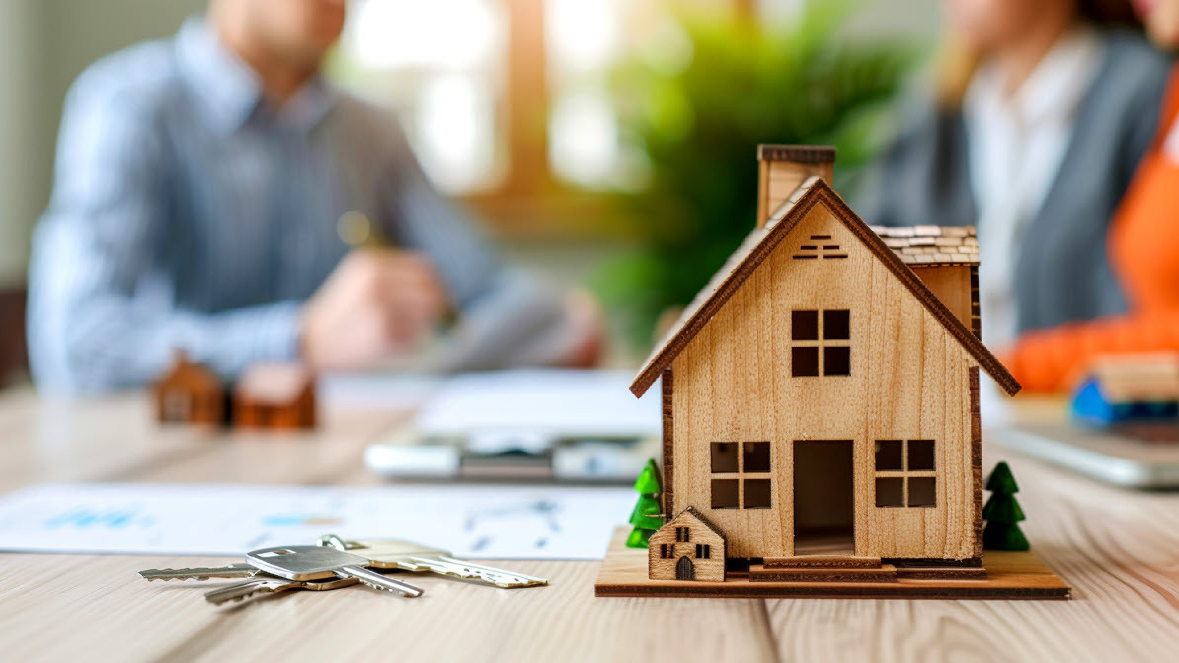 Sulla scrivania c’è una casa unifamiliare modello, accanto ci sono le chiavi di casa e sullo sfondo tre persone che partecipano a una riunione.
