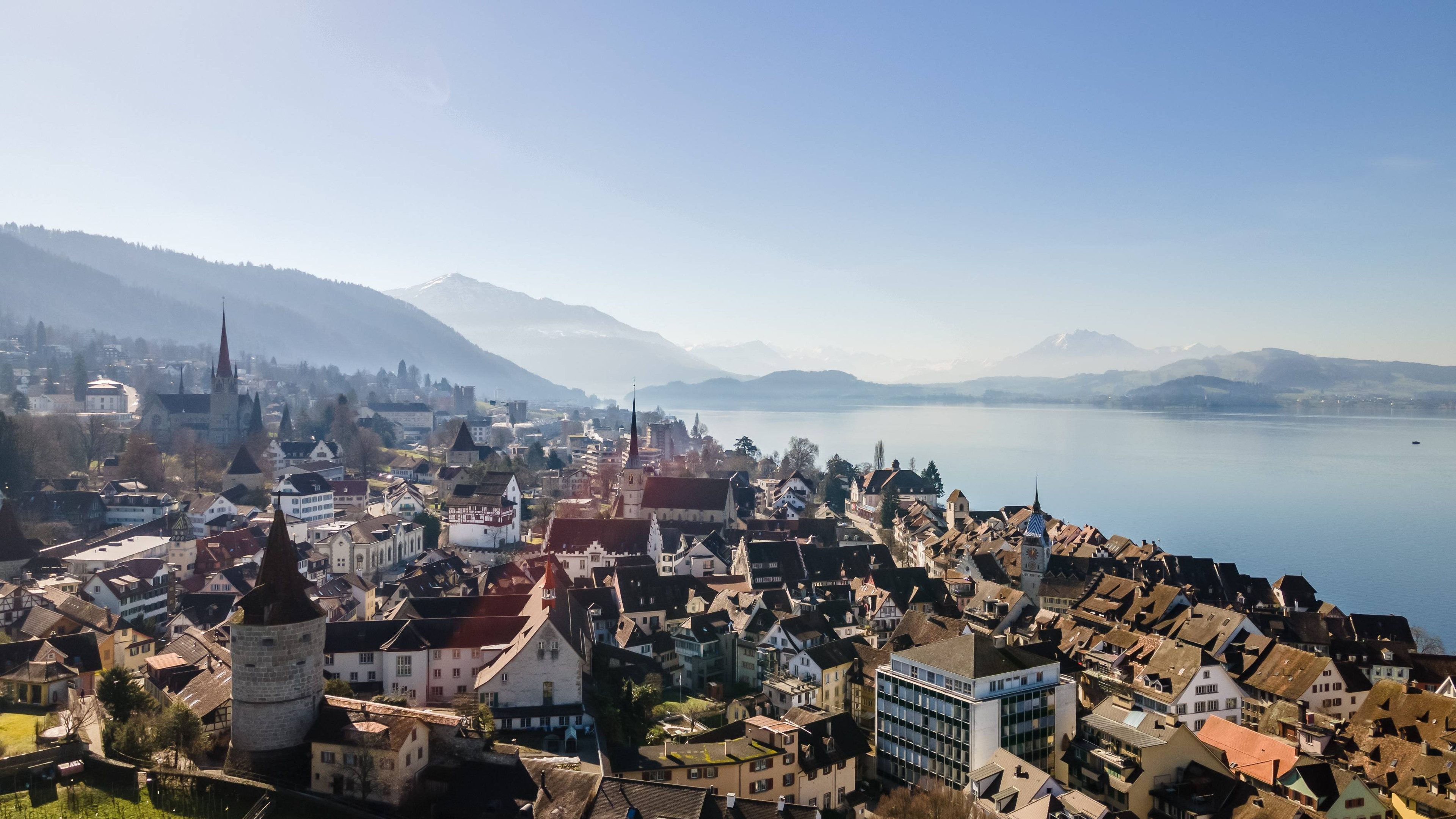 Immagine da drone della città di Zugo e del Lago di Zugo. Il tempo è autunnale: soleggiato e nebbia. Sullo sfondo si vedono le montagne.