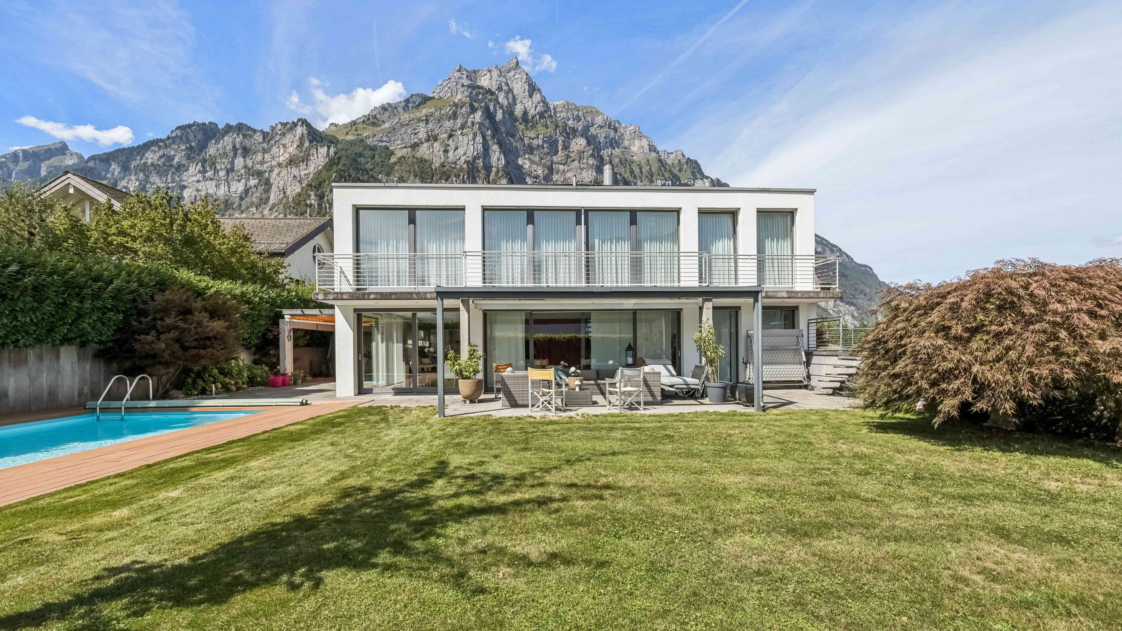 Luminosa casa unifamiliare con tetto piano con balcone e posto a sedere. Nel giardino c’è una piscina, un’ampia area verde e arbusti. Sullo sfondo si vedono le montagne.