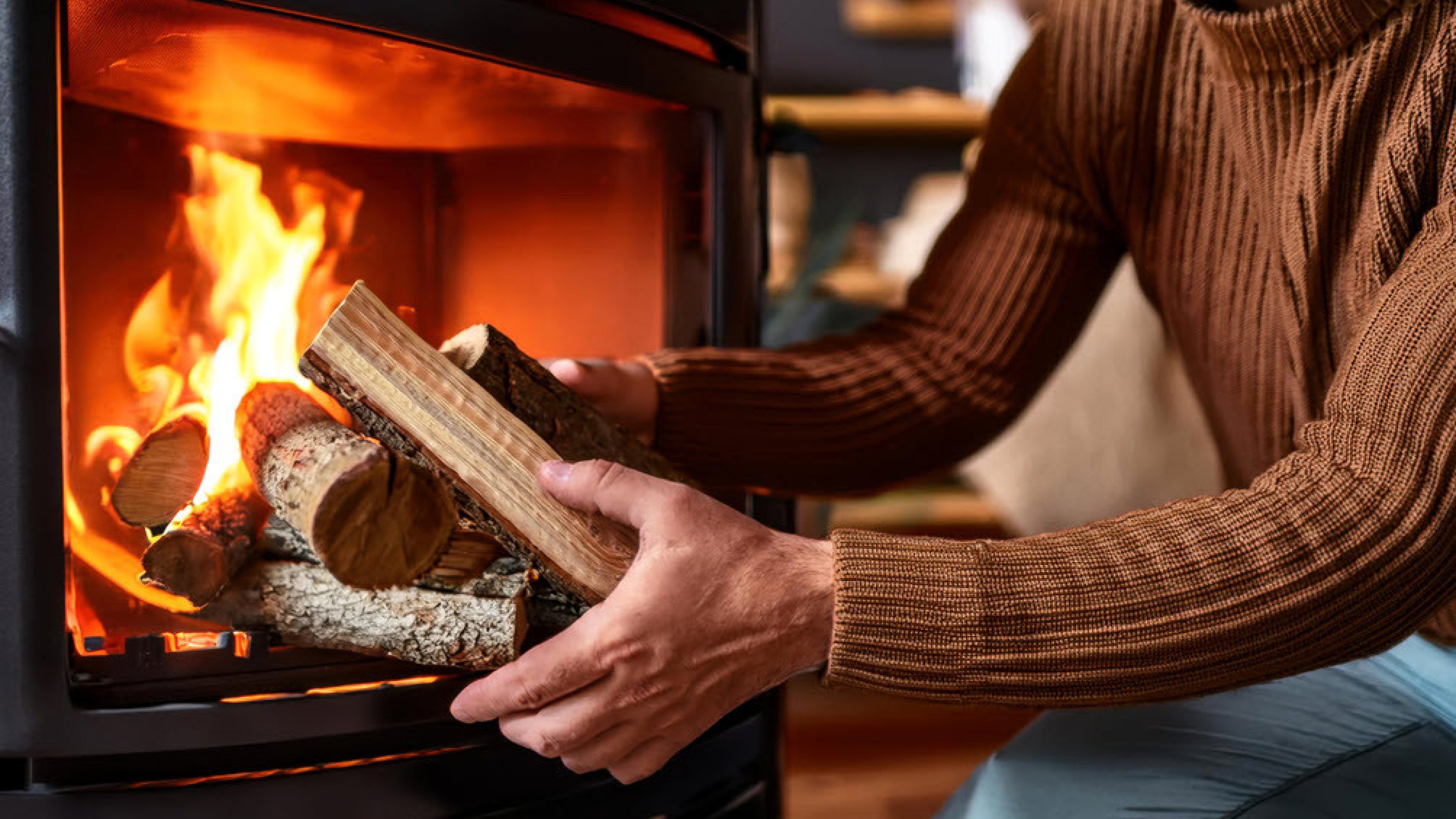 Il fuoco brucia nella stufa svedese. Un uomo con un maglione marrone mette nuova legna nella stufa.