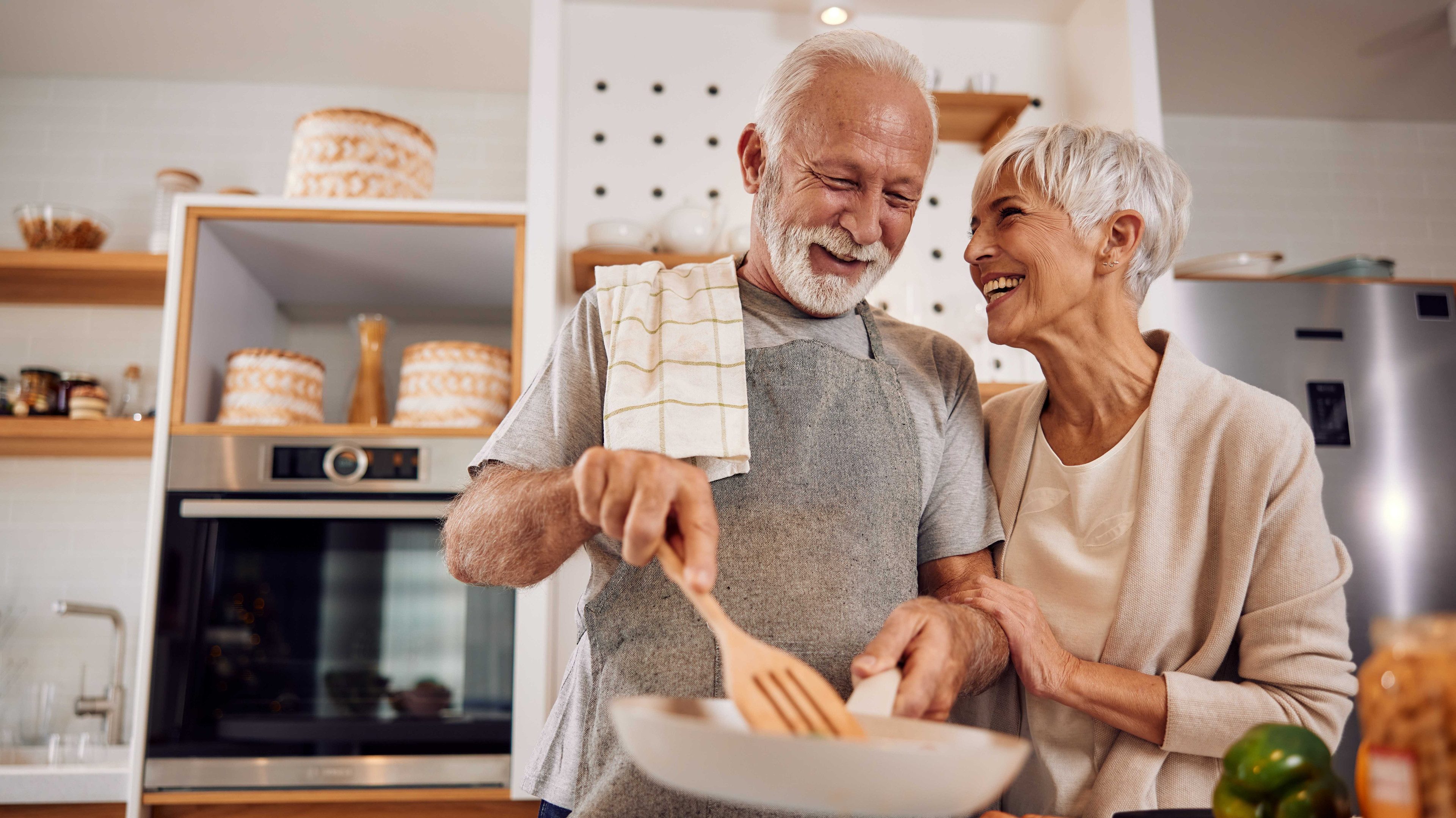 Un signore e una donna anziana stanno cucinando qualcosa insieme in cucina. Entrambi ridono e sono allegri.