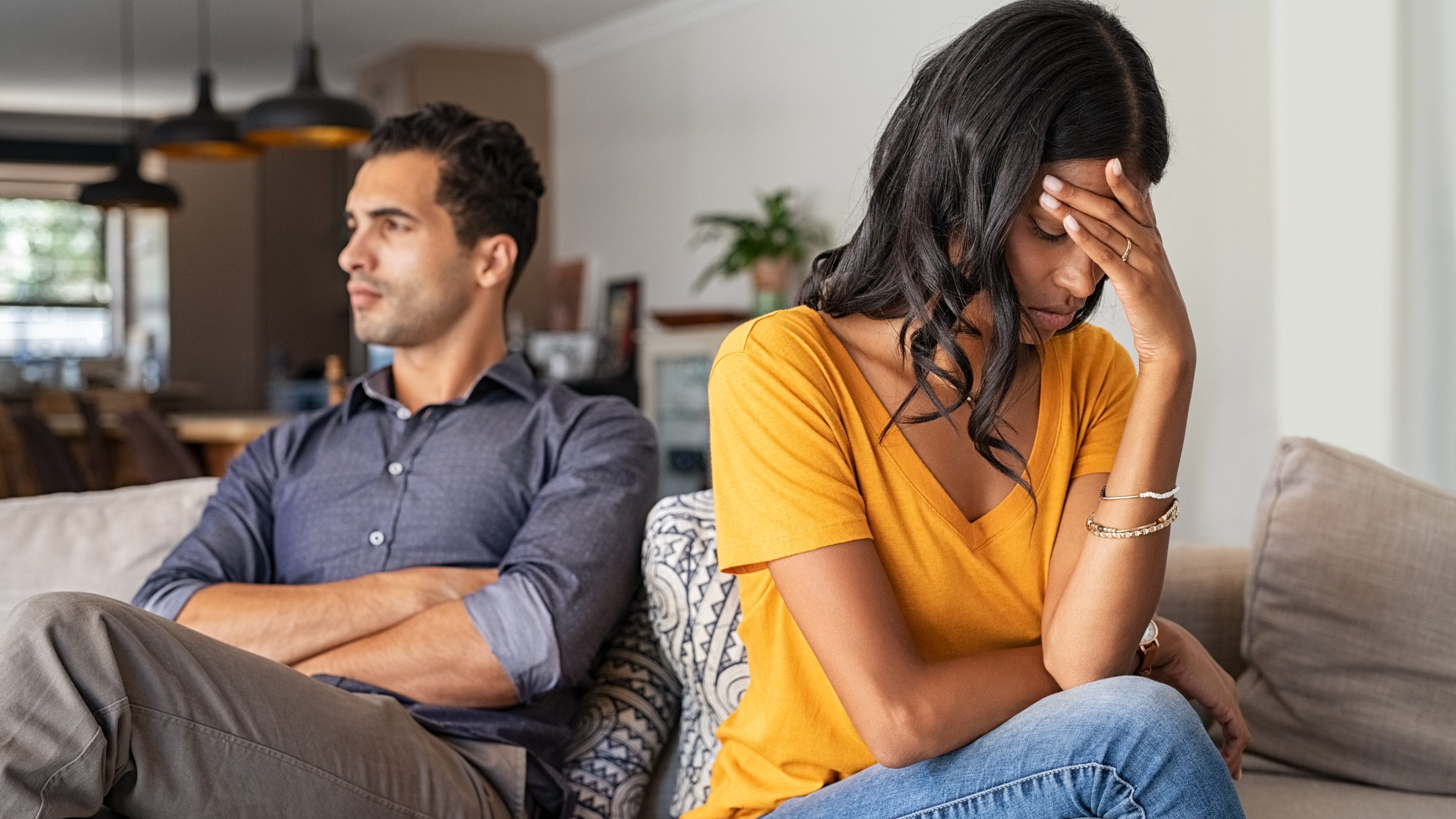 Middle eastern young couple sitting on couch after a fight. Sad indian woman sitting with hand on head after quarrel with boyfriend at home. Angry latin couple ignoring each other on the sofa, having relationship troubles.