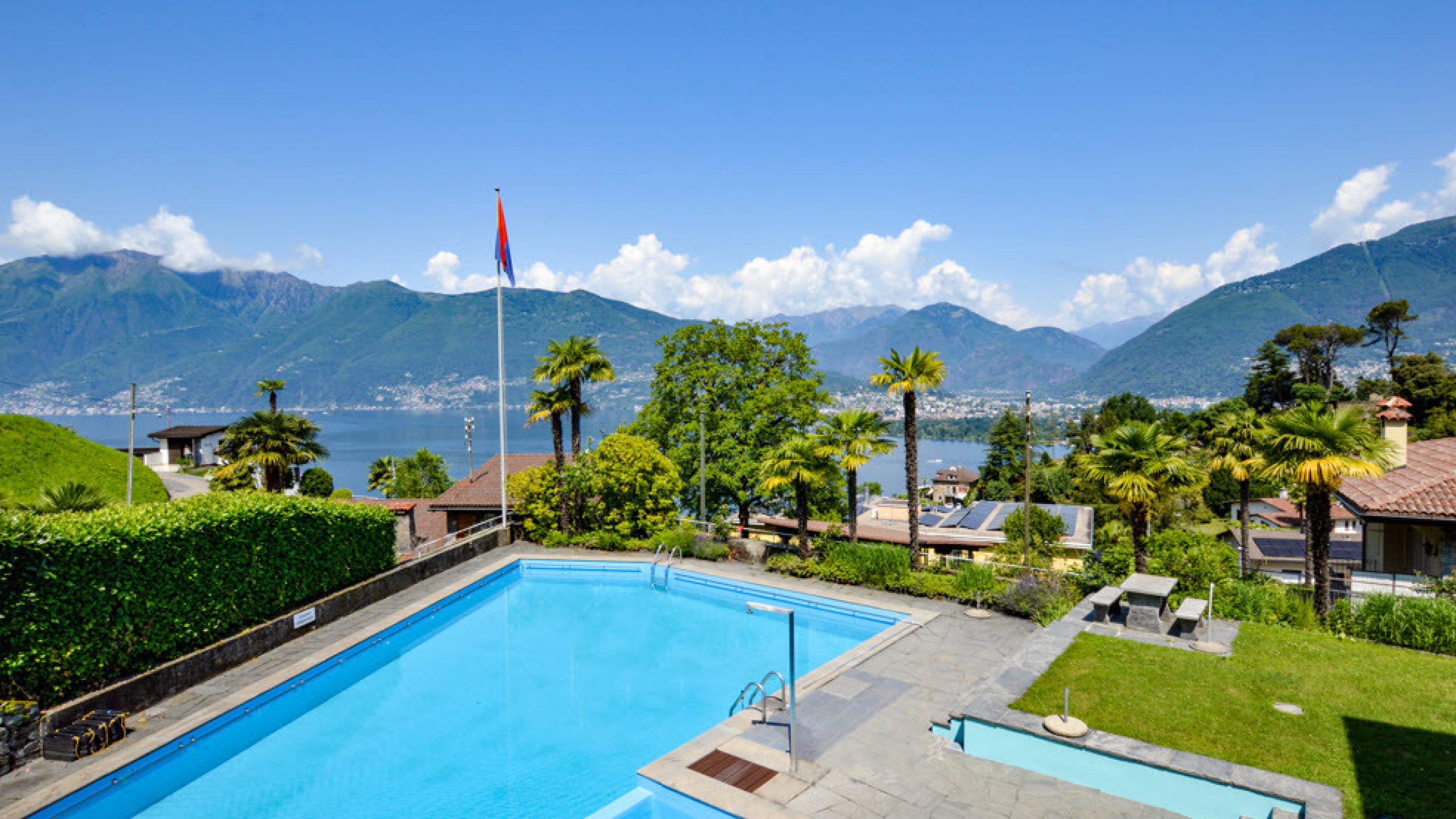 Vista aerea: Giardino con palme e grande piscina, sullo sfondo un lago e un paesaggio montano