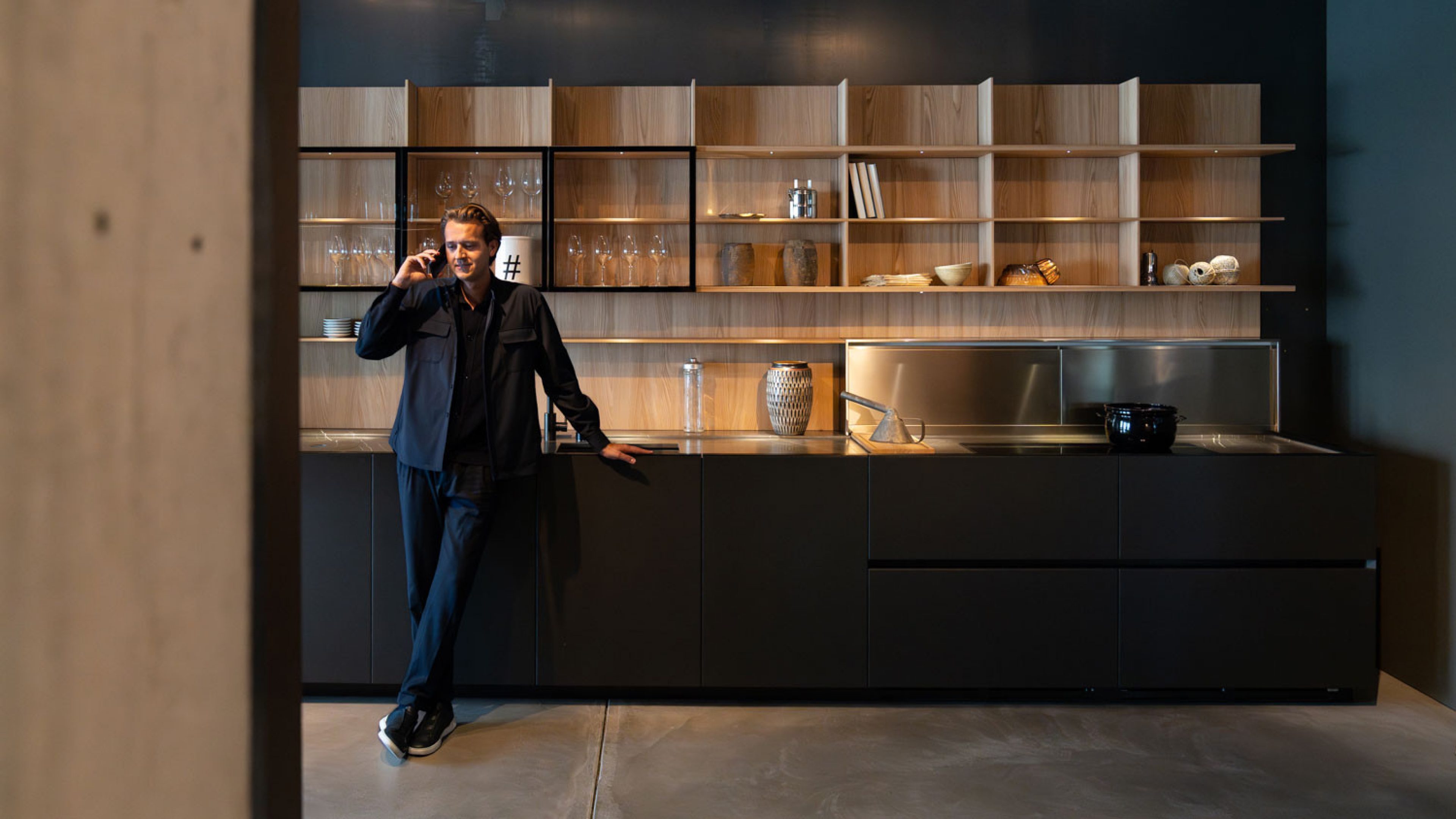 Un uomo è in una cucina moderna e minimalista con armadi scuri e scaffali di legno aperti, indossa un abito scuro e con una mano si appoggia rilassato all’isola della cucina mentre parla al telefono.