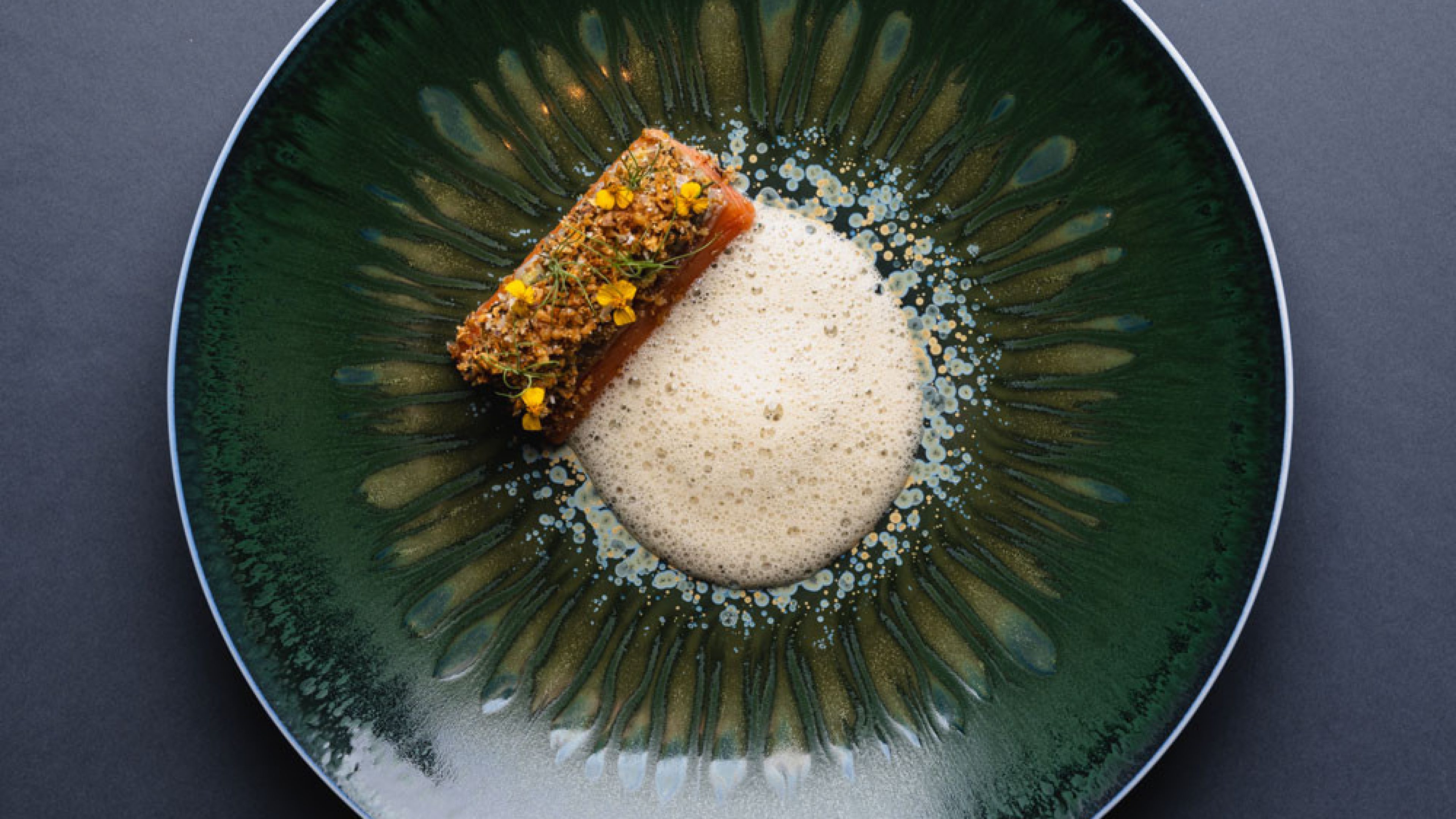 Una pietanza su di un piatto rotondo di colore verde scuro con un elemento schiumoso chiaro e un ingrediente rettangolare accanto su cui sono disposte erbe aromatiche. 