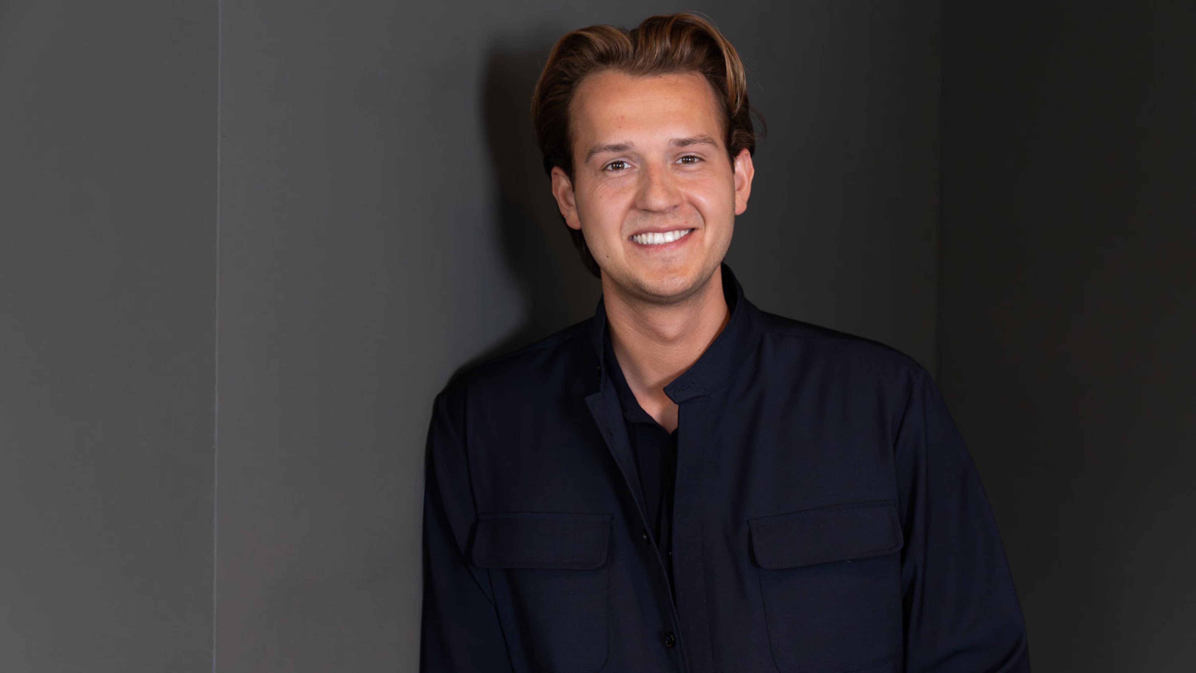 Ritratto di un giovane uomo coi capelli castani e un bel sorriso su sfondo scuro. Indossa un outfit nero in tinta unita.