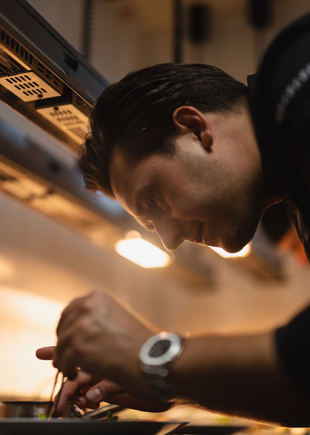 Primo piano di un cuoco concentrato con i capelli pettinati all’indietro in una cucina professionale. Si piega su un piatto e sistema il piatto con precisione. Una luce calda illumina il suo viso e la sua mano.