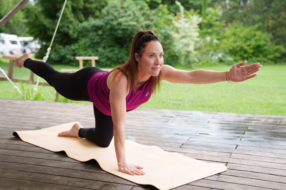Una donna con maglia sportiva viola e leggins neri è in equilibrio sul ginocchio sinistro e sul palmo della mano destra sopra un tappetino da yoga beige. Allunga il braccio sinistro dritto in avanti e guarda davanti a sé, mentre la gamba destra è rivolta dritta all’indietro.