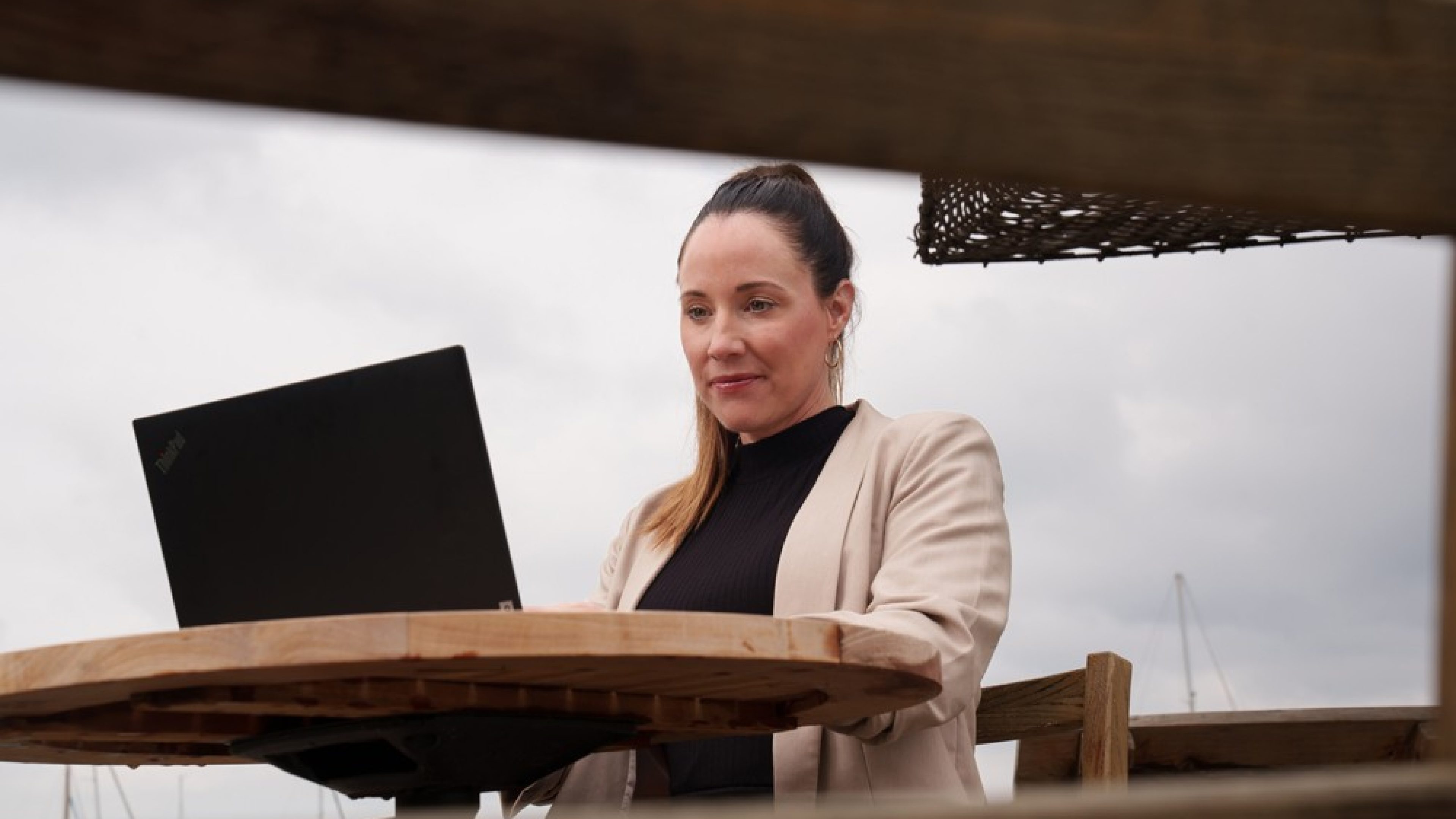 Una donna con un blazer beige e un maglione nero siede concentrata davanti al laptop all’aperto.