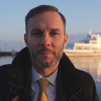 Uomo con cravatta guarda nell’obiettivo della telecamera. Dietro di lui si vede un battello che attracca.