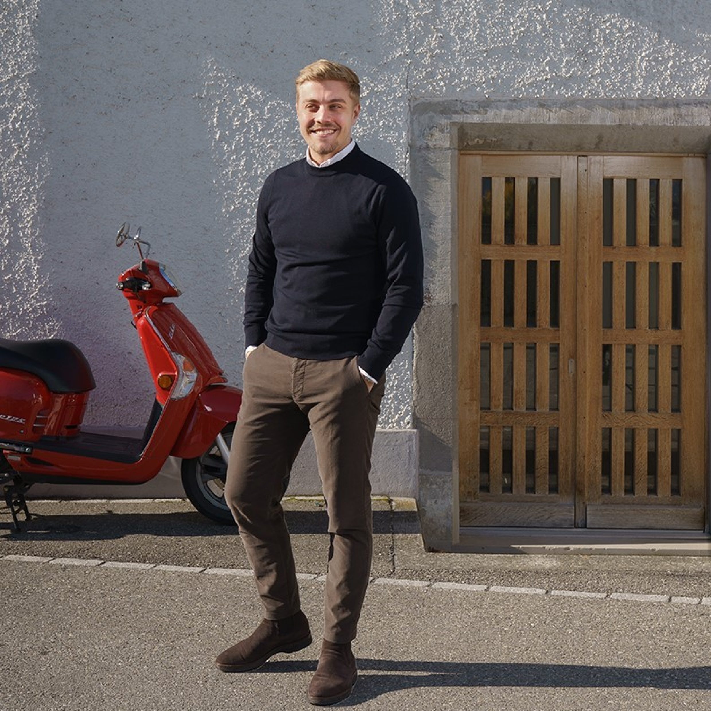 Un uomo si trova davanti a un edificio bianco e tiene le mani in tasca. Dietro l’uomo, a sinistra, c’è una Vespa rossa, e a destra, una porta di legno che conduce all’edificio bianco.