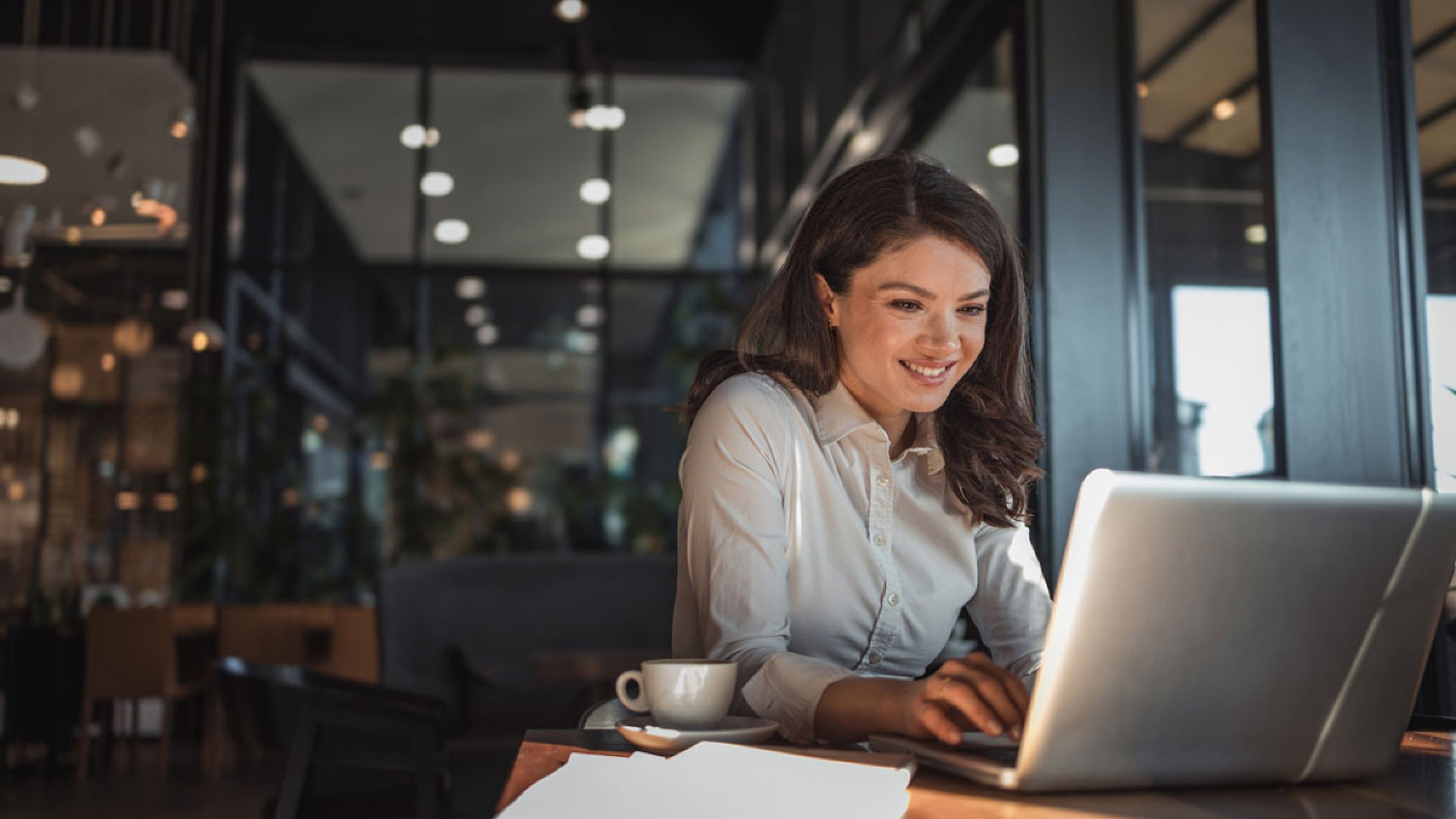 Una giovane donna siede in un moderno caffè, sorride e lavora concentrata al suo laptop. Davanti a lei, sul tavolo, ci sono una tazza di caffè e alcuni fogli di carta bianchi. Il locale è ben illuminato, con un’atmosfera elegante e grandi finestre che inondano l’ambiente di luce naturale. 