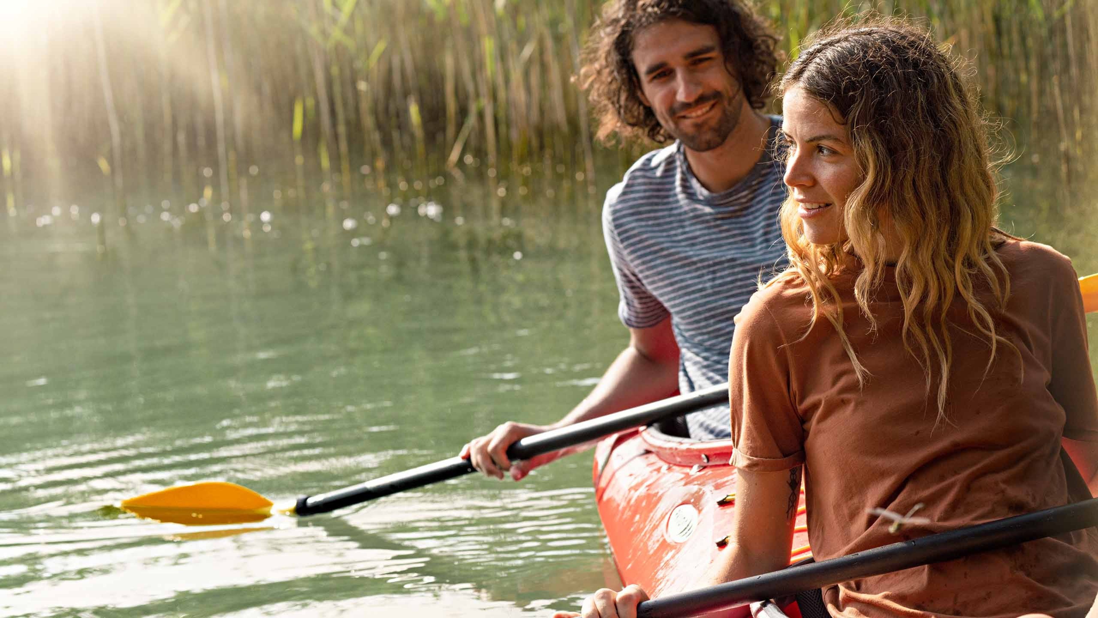 Una donna e un uomo vanno in canoa sul lago. Sullo sfondo si vedono delle canne. C’è il sole.