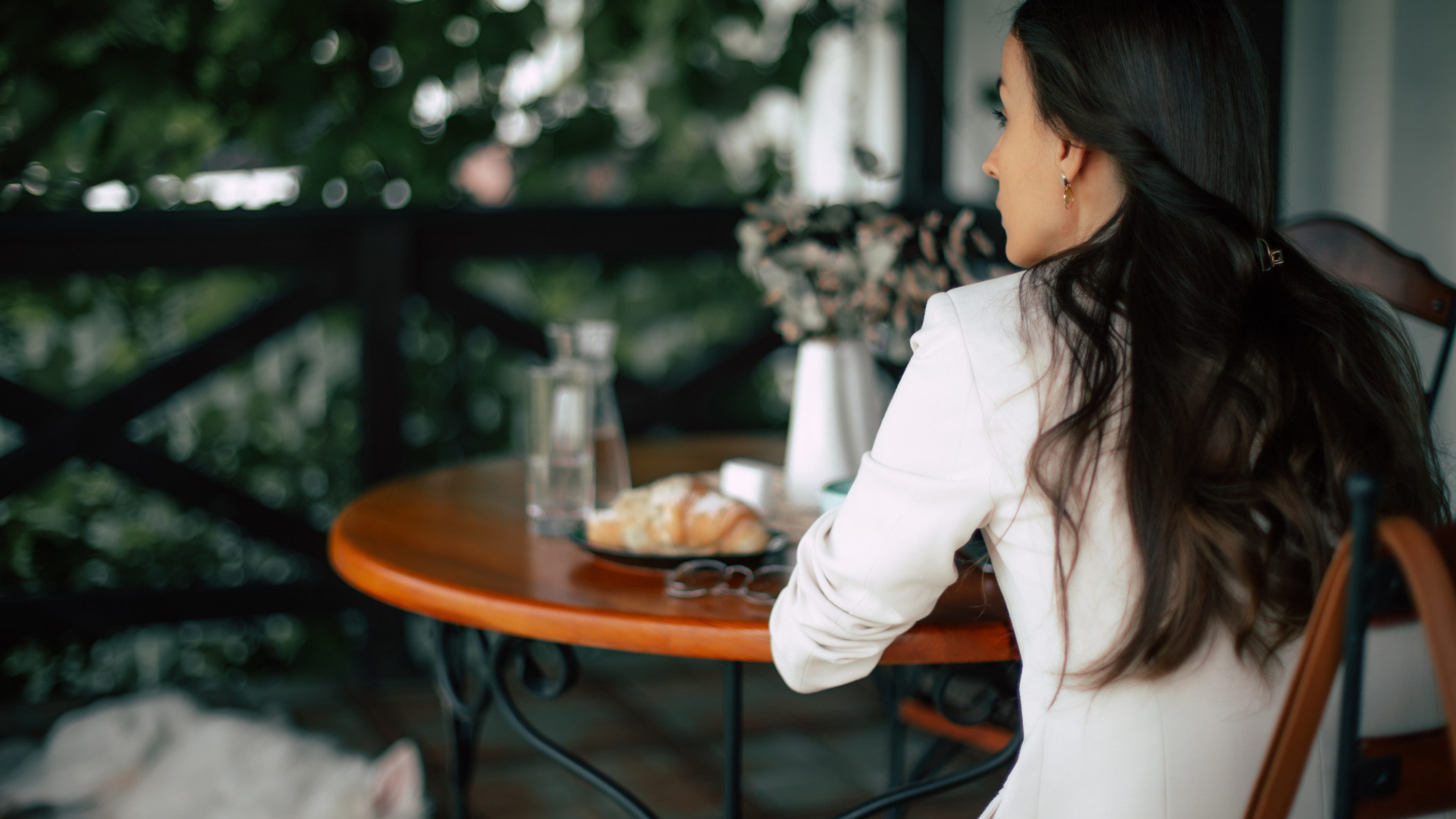 Una donna con i capelli lunghi e scuri siede su una terrazza, dando la schiena alla fotocamera, a un tavolo rotondo di legno. Davanti a lei un bicchiere d’acqua, una caraffa e un croissant. Sullo sfondo si vedono piante verdi e una ringhiera, con accanto un cane sdraiato a terra.