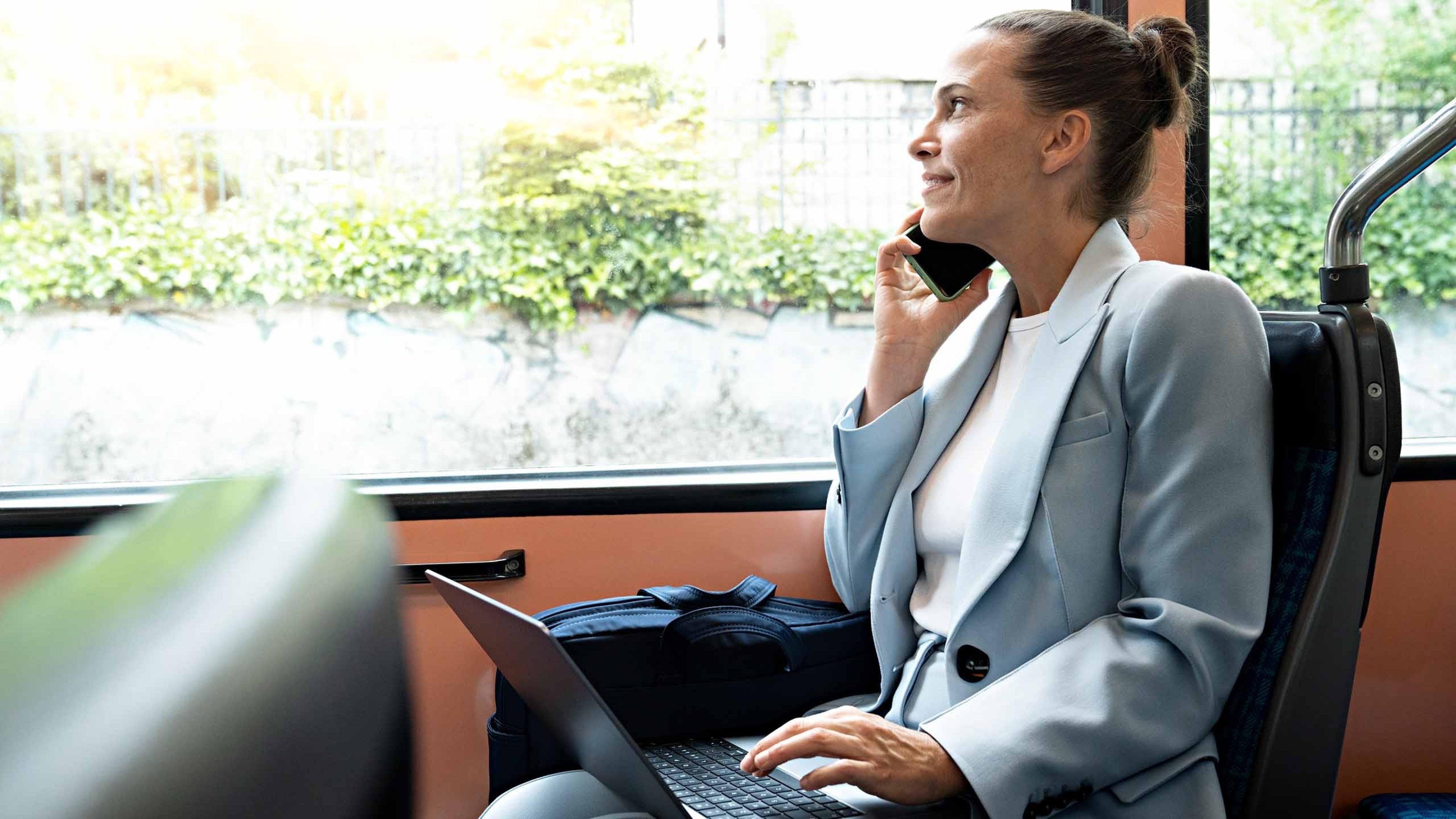 Una donna è seduta in tram con il laptop sulle ginocchia e telefona mentre guarda fuori dal finestrino.