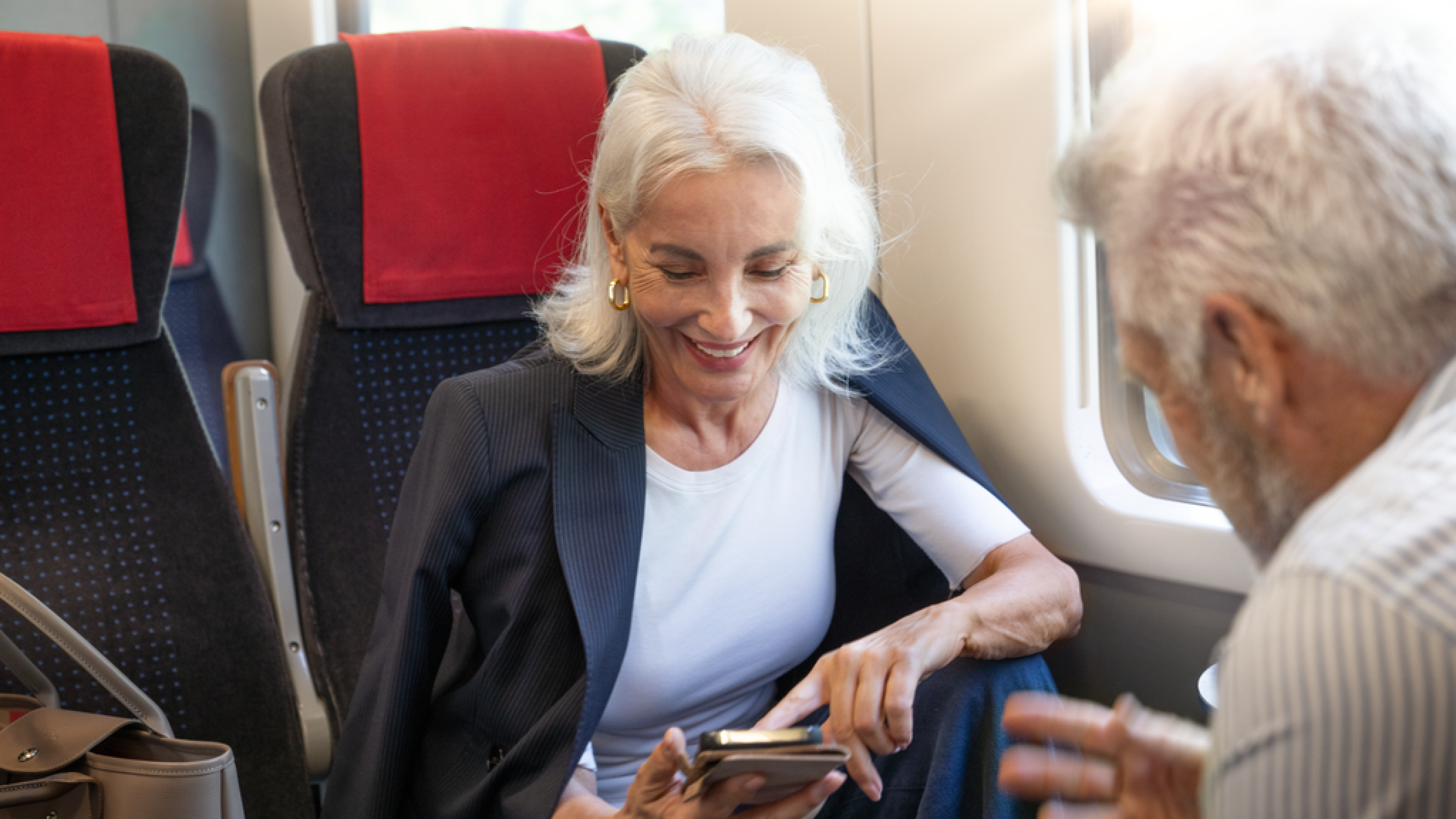 Una donna anziana è seduta in treno e guarda sorridente il suo smartphone.