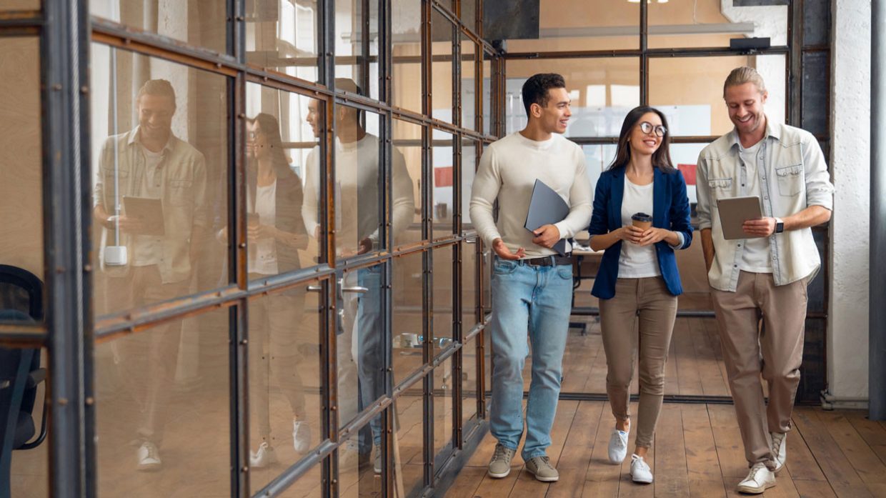 Tre giovani professionisti camminano insieme in un ufficio moderno con pareti di vetro. Sembrano motivati e impegnati in una conversazione mentre tengono in mano laptop e tablet.